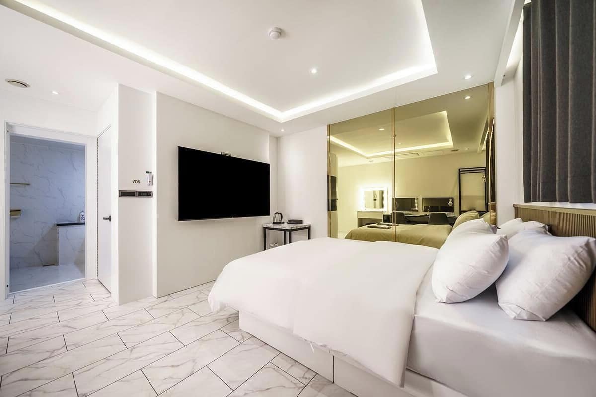 Una camera d'albergo moderna con un letto grande, una televisione a schermo piatto fissata al muro, un piccolo comodino e un bagno visibile attraverso una porta aperta.