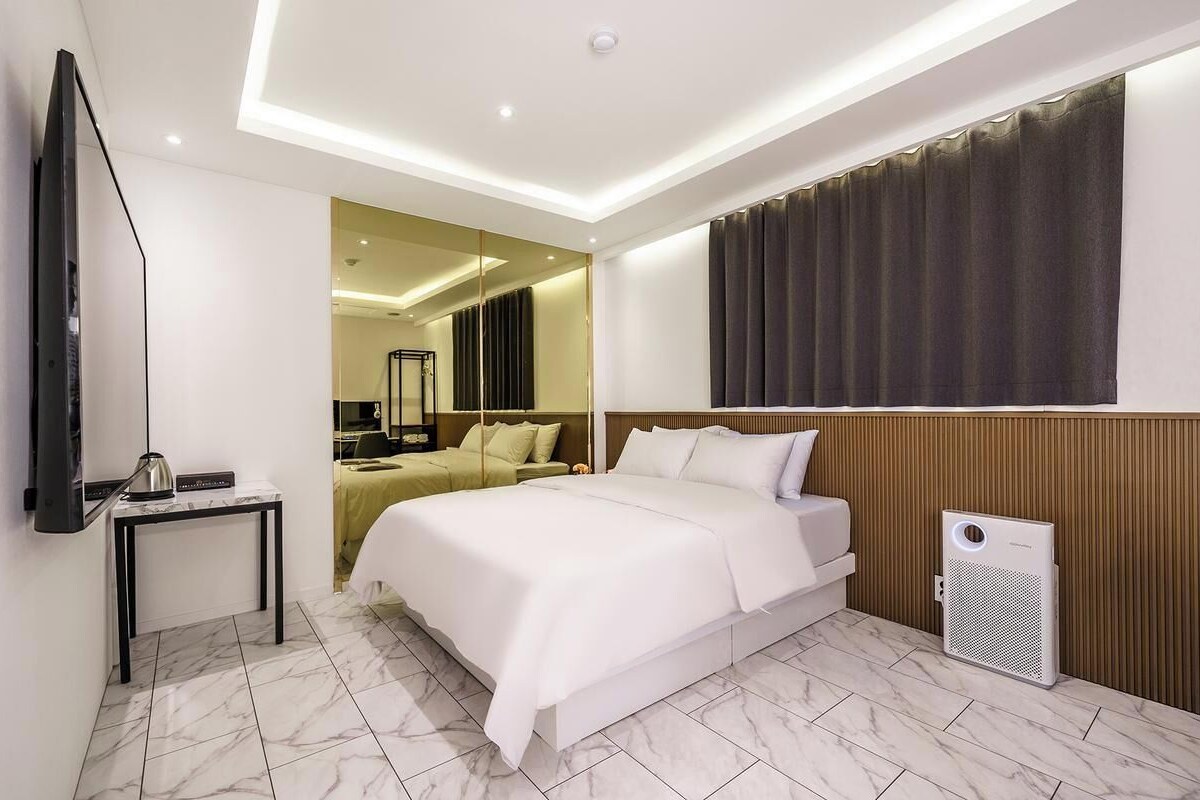 A hotel room with a large bed, a bedside table with a lamp, a flat-screen TV, and a wall-mounted air conditioning unit.