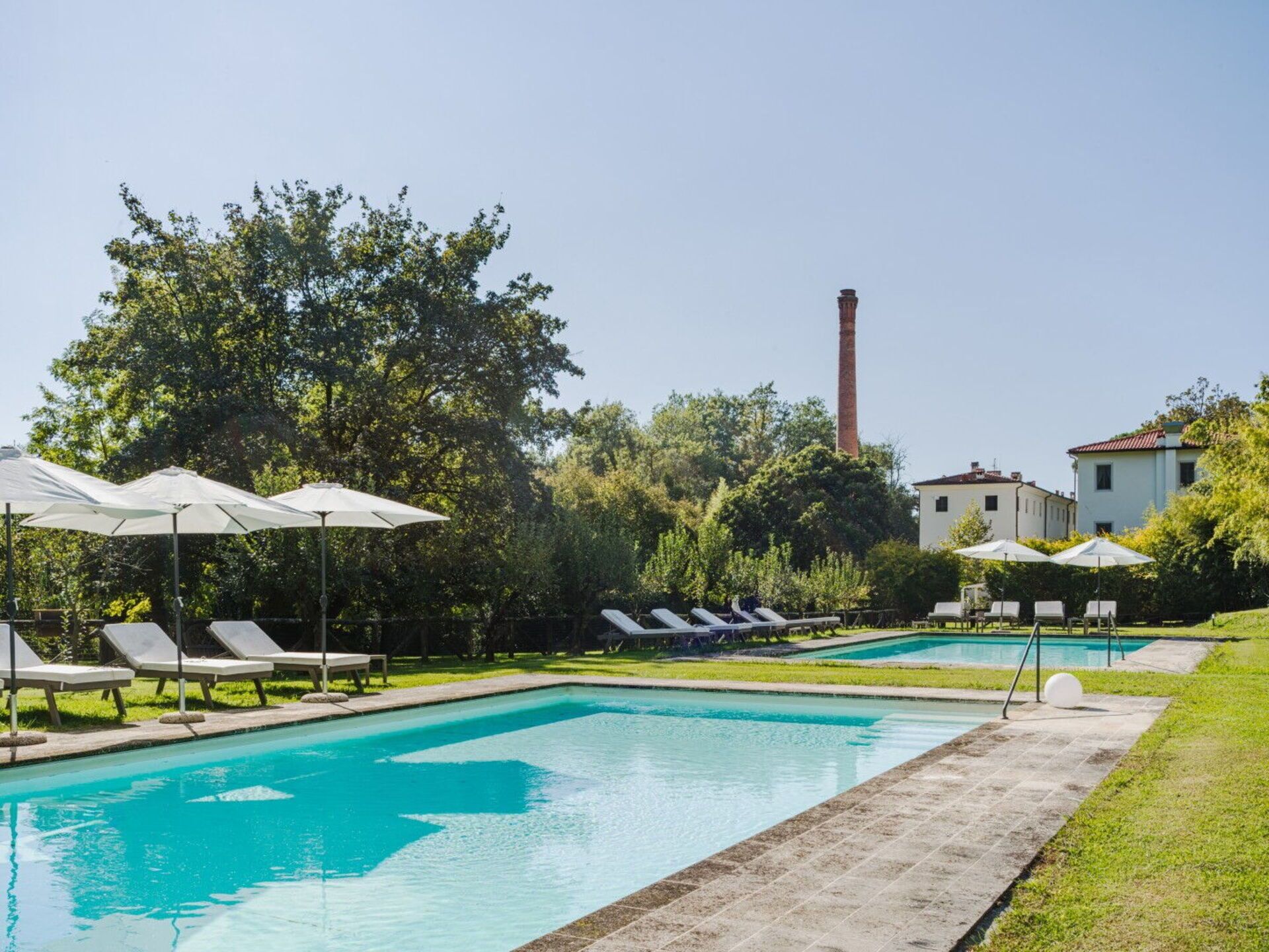 A pool area with lounge chairs and umbrellas, surrounded by greenery and a building with a chimney.