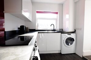 A compact kitchen with a washing machine, a coffee maker, and a window with pink blinds. - Gloucester-4 Bed Home for Work & Leisure (Gloucester)