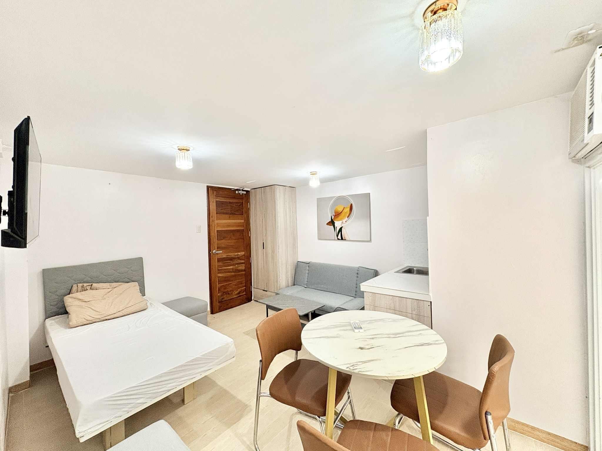 A compact hotel room with a bed, a sofa, a small dining table with chairs, a kitchen area, and a door.