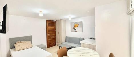 A compact hotel room with a bed, a sofa, a small dining table with chairs, a kitchen area, and a door.