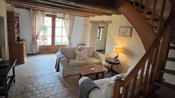 Un salon chaleureux avec un sol en pierre, un escalier en bois et un canapé agrémenté d’un coussin.