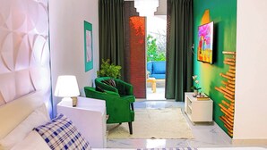 A hotel room with a bed, a green armchair, a white nightstand, a lamp, a TV on the wall, and a view of the outside balcony. - Agapimou apartments H (Gaspar Hernandez)