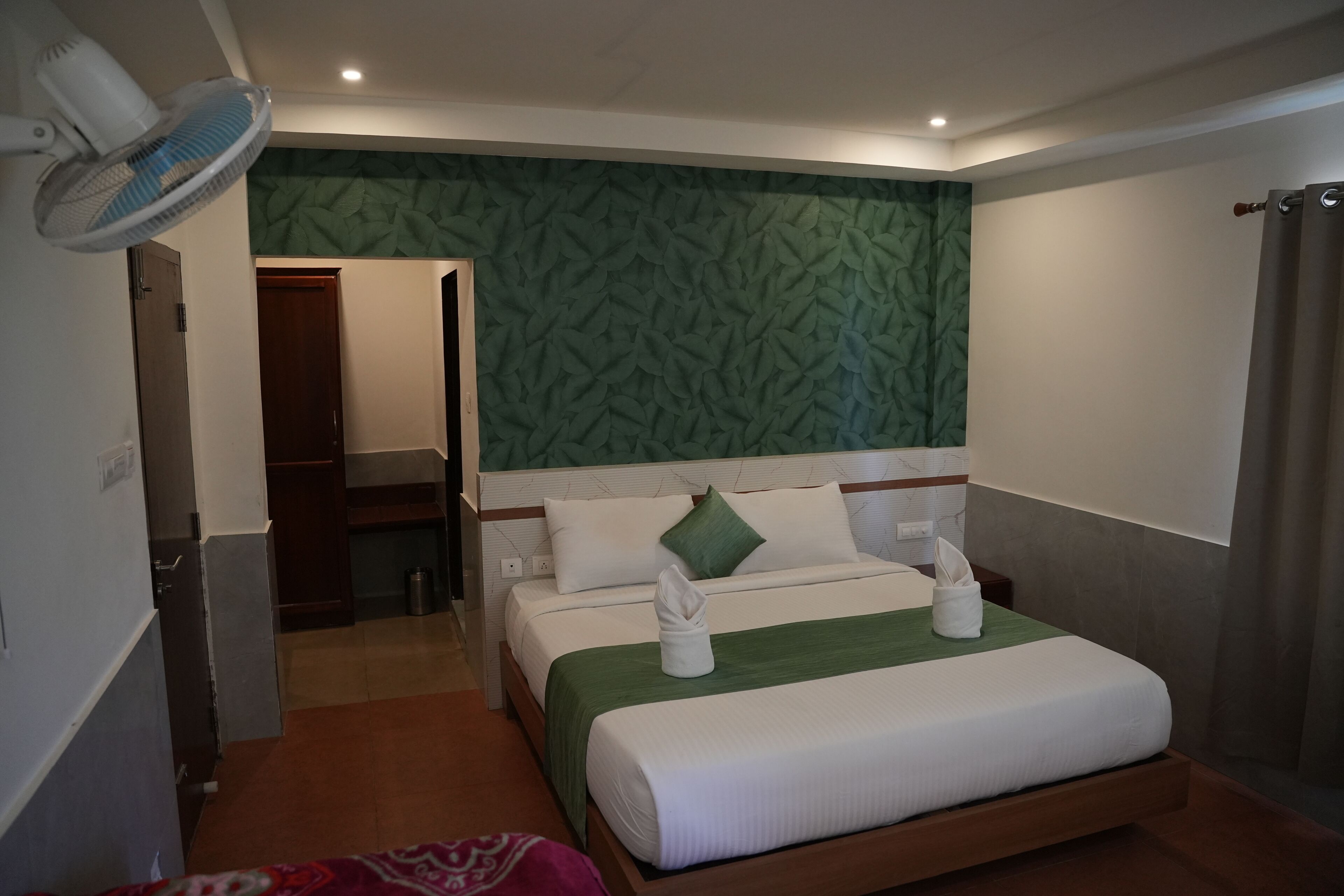 A hotel room with a bed, a fan, a green patterned wall, and a door leading to a bathroom.