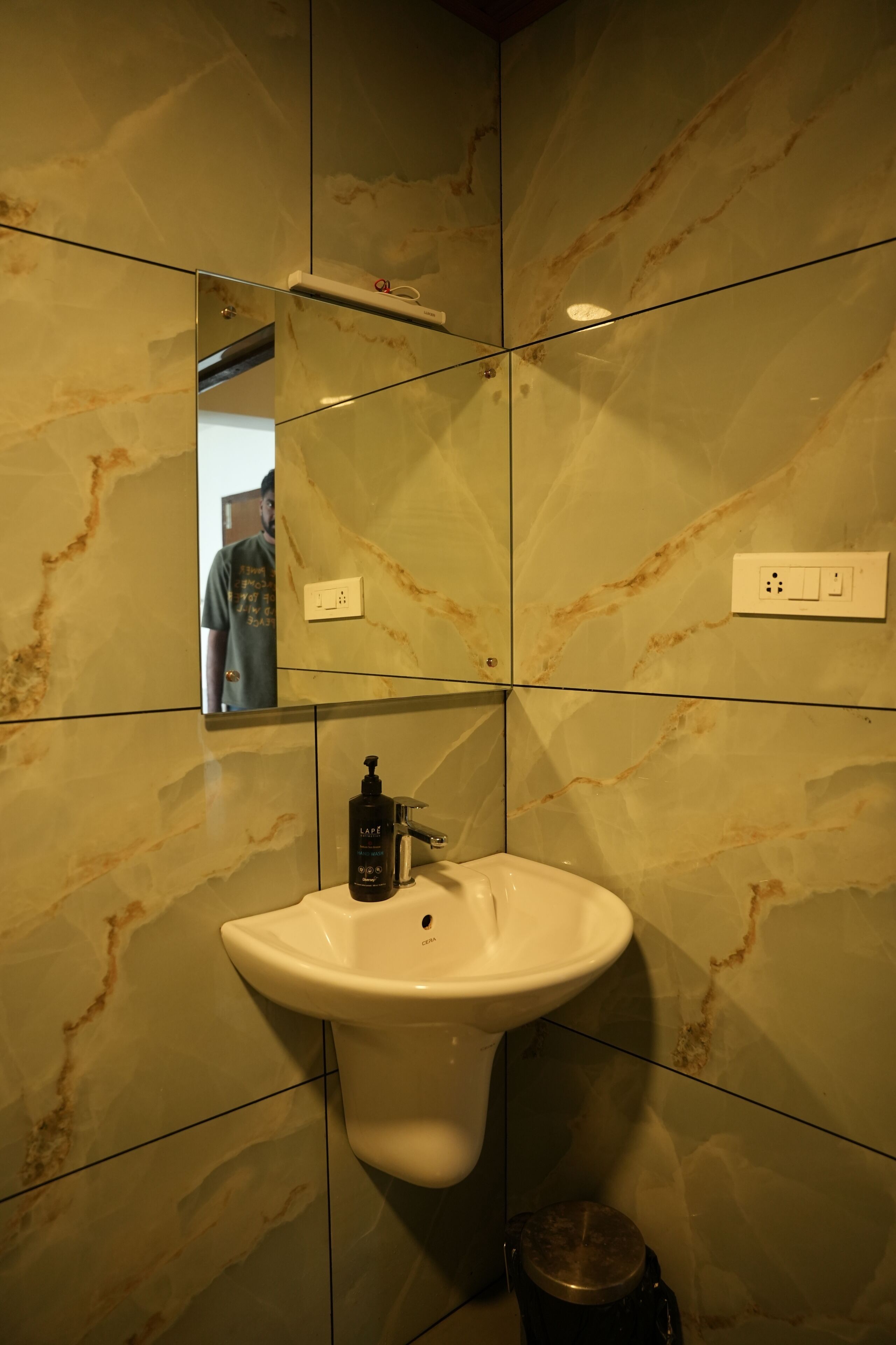 A bathroom with a marble wall, a sink, a mirror, and a trash bin.