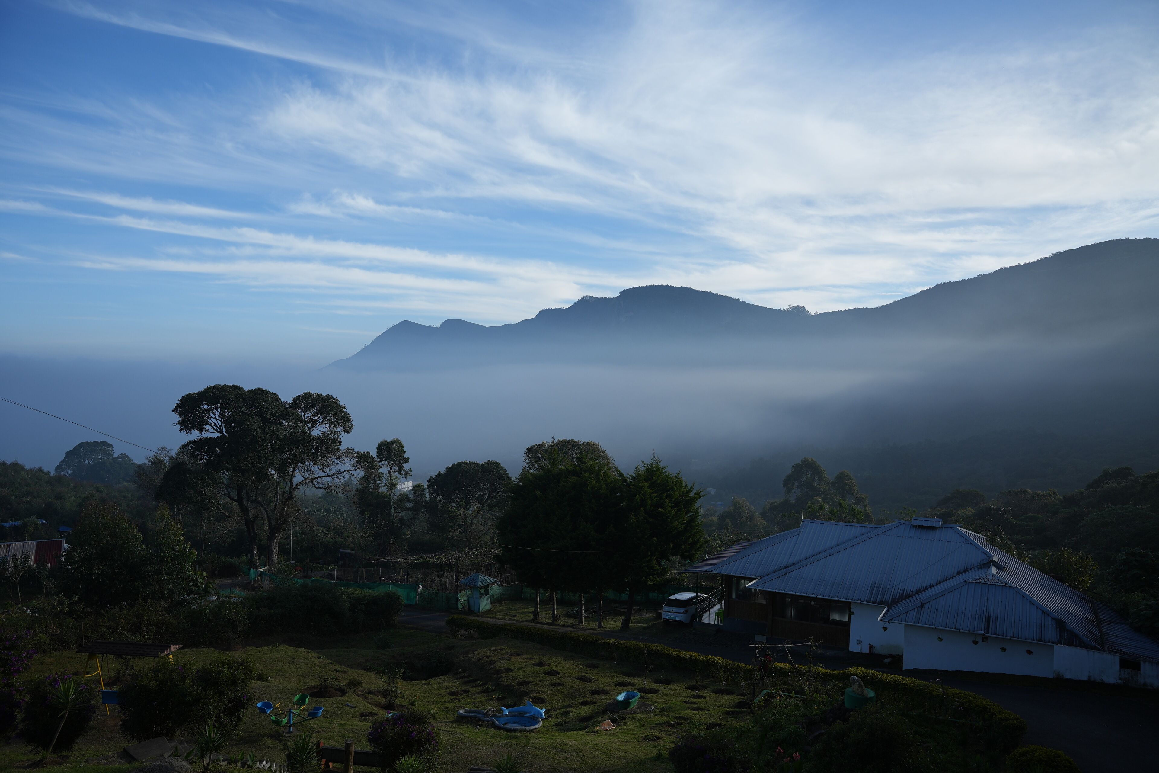 Paisaje rural con una casa, un pequeño jardín y vistas a montañas envueltas en niebla.