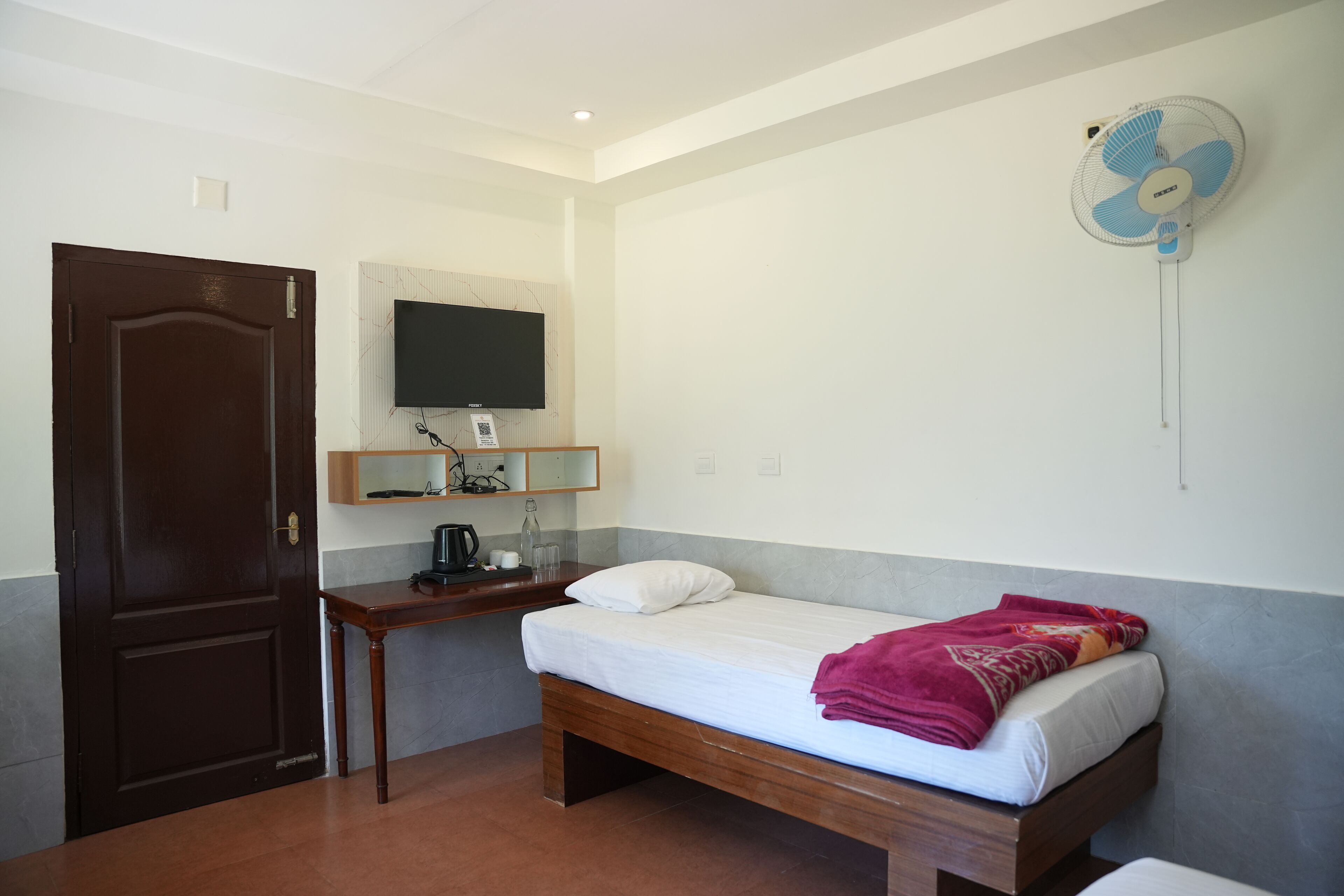 A small, clean room with a bed, a desk, a television, and a wall-mounted fan.