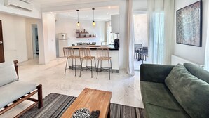 Ein modernes Wohnzimmer mit einem grünen Sofa, einem Holztisch und einem Essbereich, im Hintergrund ist eine Küche zu sehen.