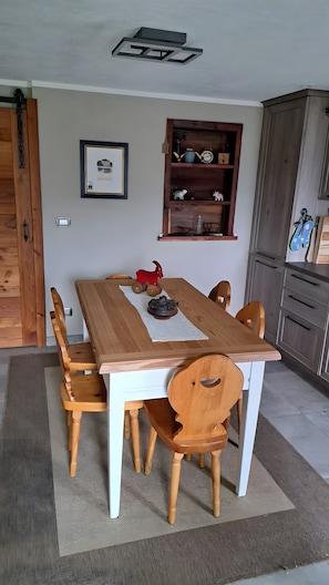 A dining area with a wooden table, chairs, a rug, a framed picture, and a wooden shelf with decorative items.