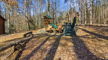 A wooden play structure with a slide and a picnic table in a forested area.