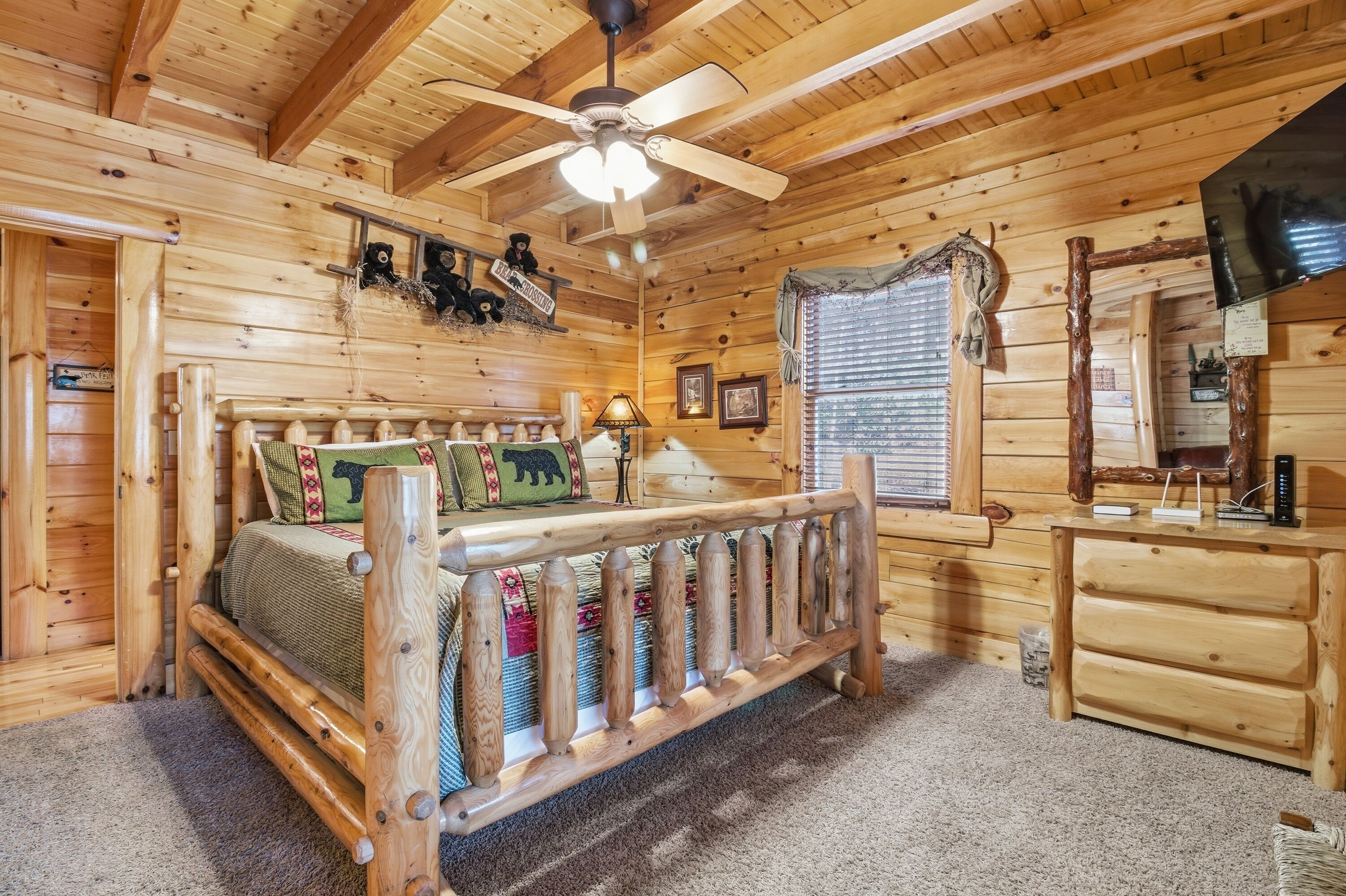 A wooden cabin bedroom with a bed, a wooden bench, a ceiling fan, a dresser, and a television.