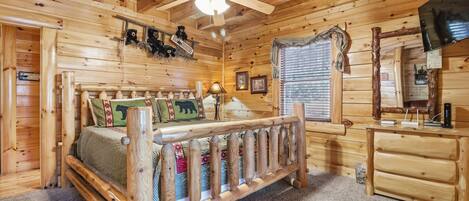 A wooden cabin bedroom with a bed, a wooden bench, a ceiling fan, a dresser, and a television.