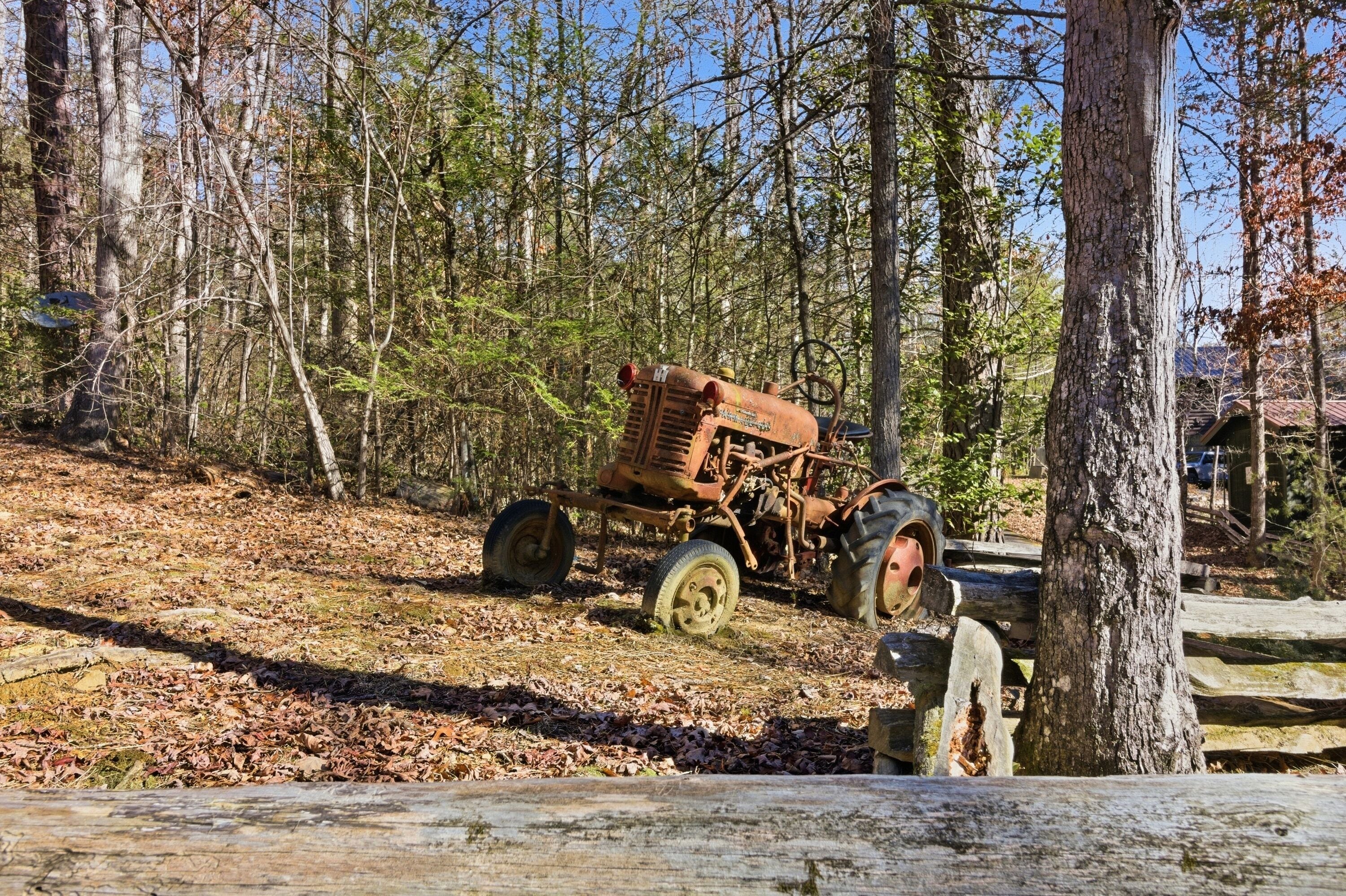 An old, rusted tractor in a forested area with trees and a wooden fence.