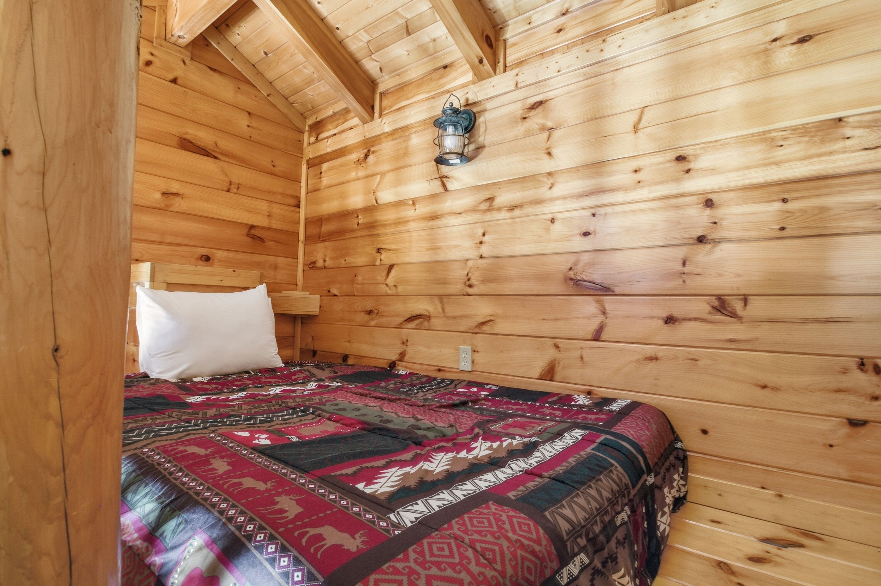 A wooden cabin room with a bed, a white pillow, and a lantern on the wall.