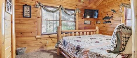 A wooden cabin room with a bed, a television, a window with curtains, and a welcome sign.