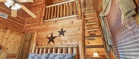 A wooden cabin room with a bed, a ceiling fan, a star decoration, and a window with blinds.