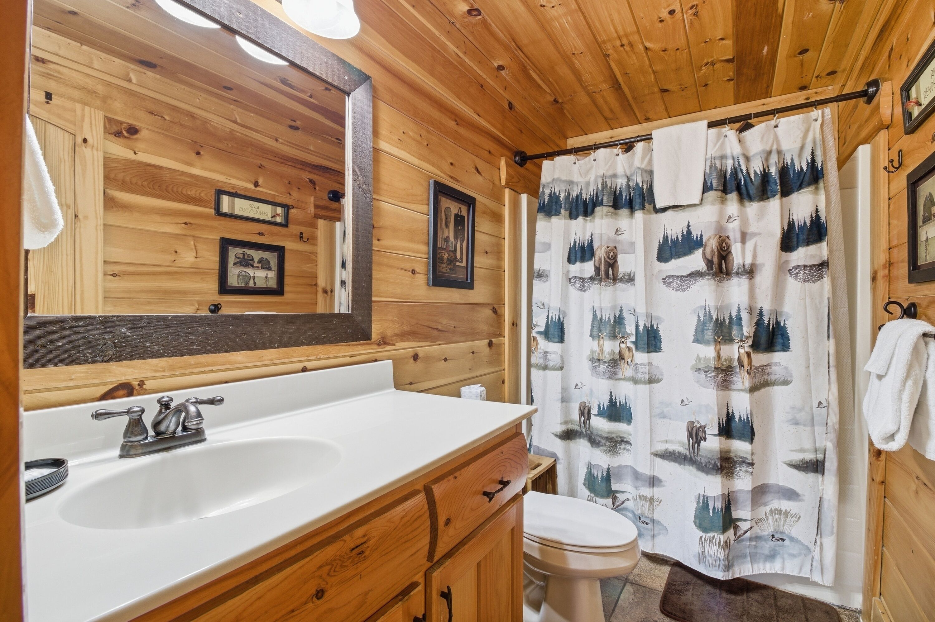A wooden cabin bathroom with a sink, toilet, and a shower curtain with a forest and wildlife print.