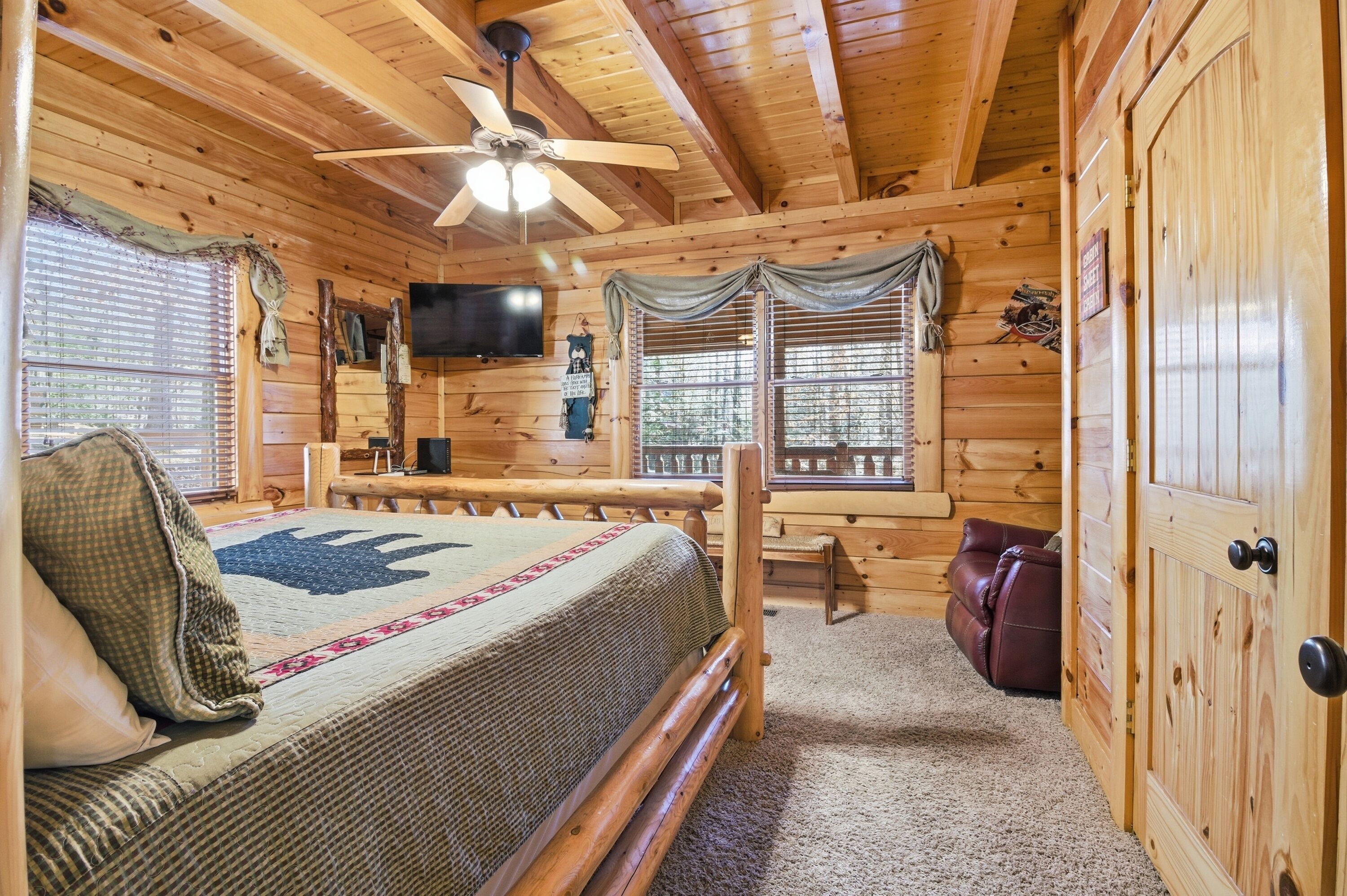 Un dormitorio de cabaña de madera con cama, ventilador de techo, televisor y ventana con cortinas.