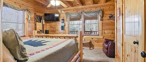 Un dormitorio de cabaña de madera con cama, ventilador de techo, televisor y ventana con cortinas.