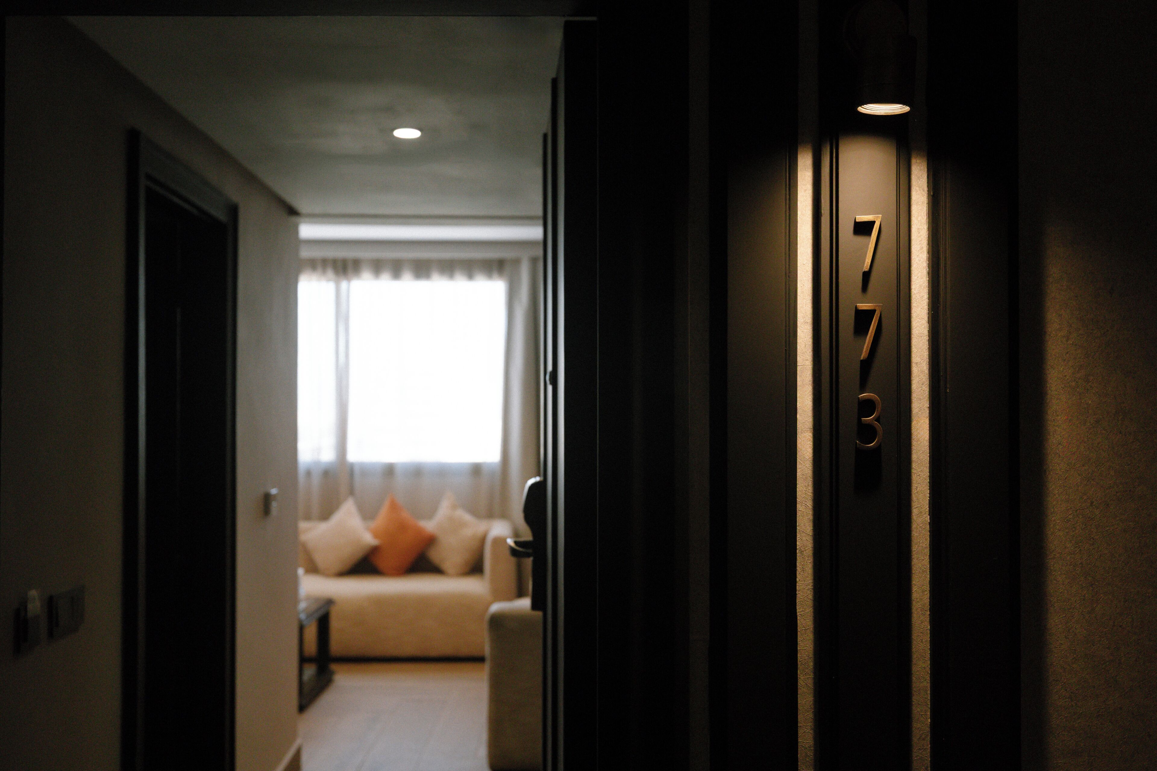 Um corredor que leva a um quarto com placa indicando '7B', um sofá com almofadas e uma janela com cortinas.