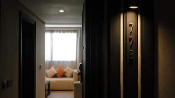 Um corredor que leva a um quarto com placa indicando '7B', um sofá com almofadas e uma janela com cortinas.