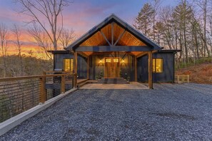 A modern log cabin with a prominent gable roof, a gravel driveway, and a wooden deck. - Riverstone Retreat | Ellijay, GA (Ellijay)