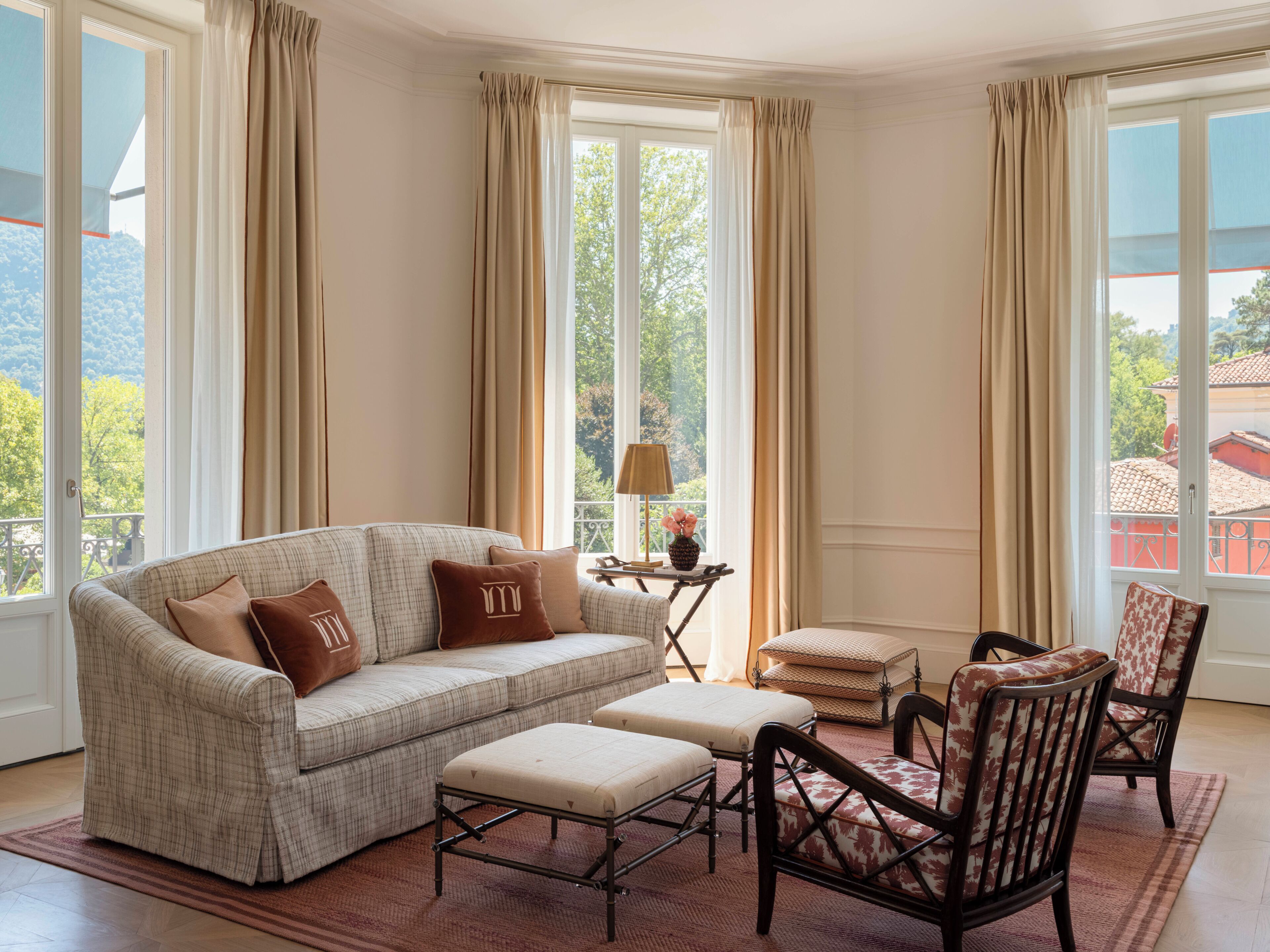 Sala de estar con sofá, reposapiés y sillas, ventanas amplias con cortinas y vistas a zonas verdes y edificios.
