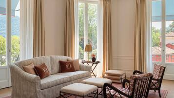 Sala de estar con sofá, reposapiés y sillas, ventanas amplias con cortinas y vistas a zonas verdes y edificios.