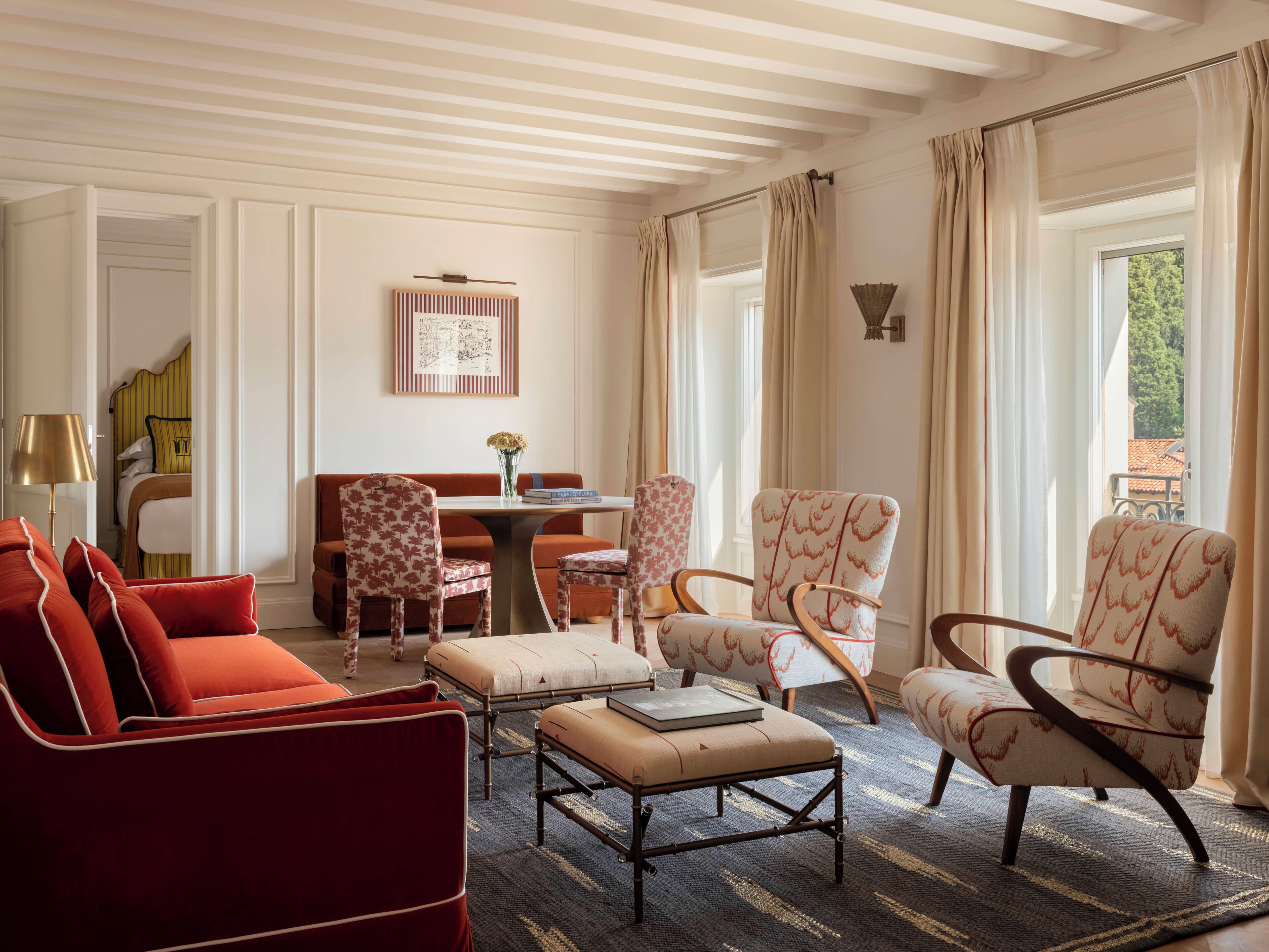 Ein helles Zimmer mit einem roten Sofa, gemusterten Sesseln, einem kleinen Tisch mit einer Vase und Blick auf Grünflächen.