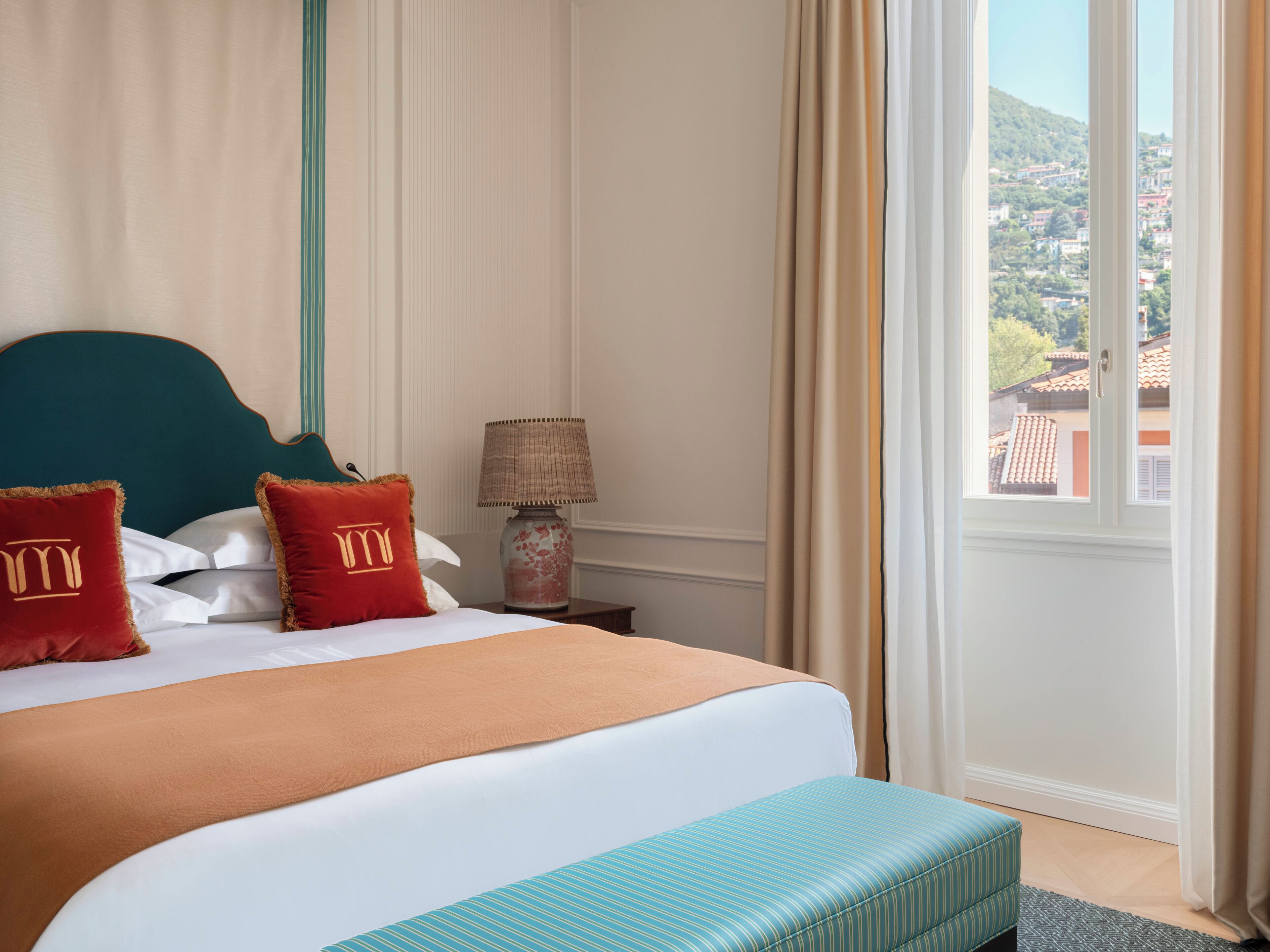 Habitación de hotel con cama, mesita de noche, lámpara y vista a un paisaje urbano por la ventana.