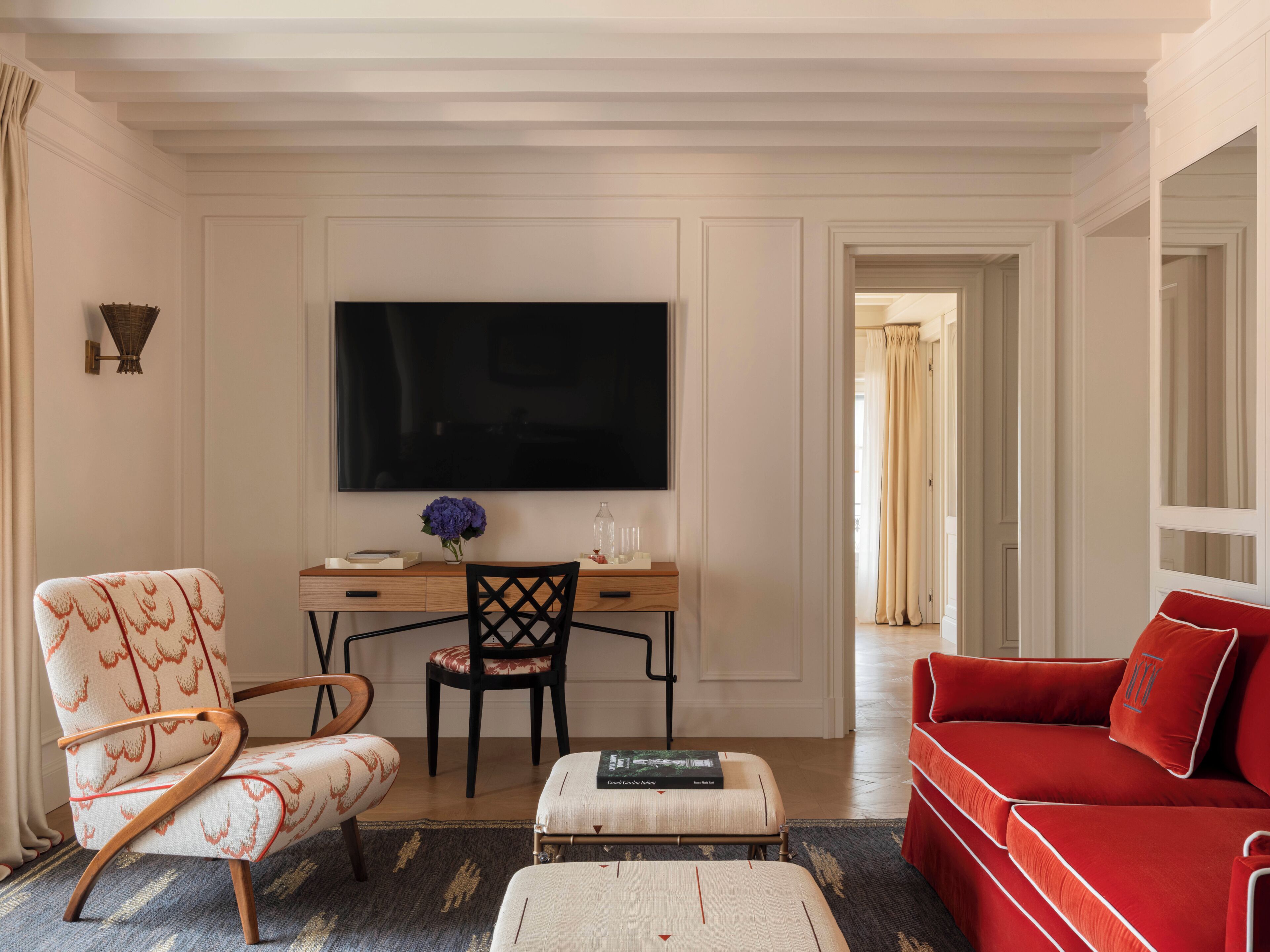 A living room with a flat-screen TV, a red sofa, a patterned armchair, a wooden coffee table, and a desk with a vase of flowers.