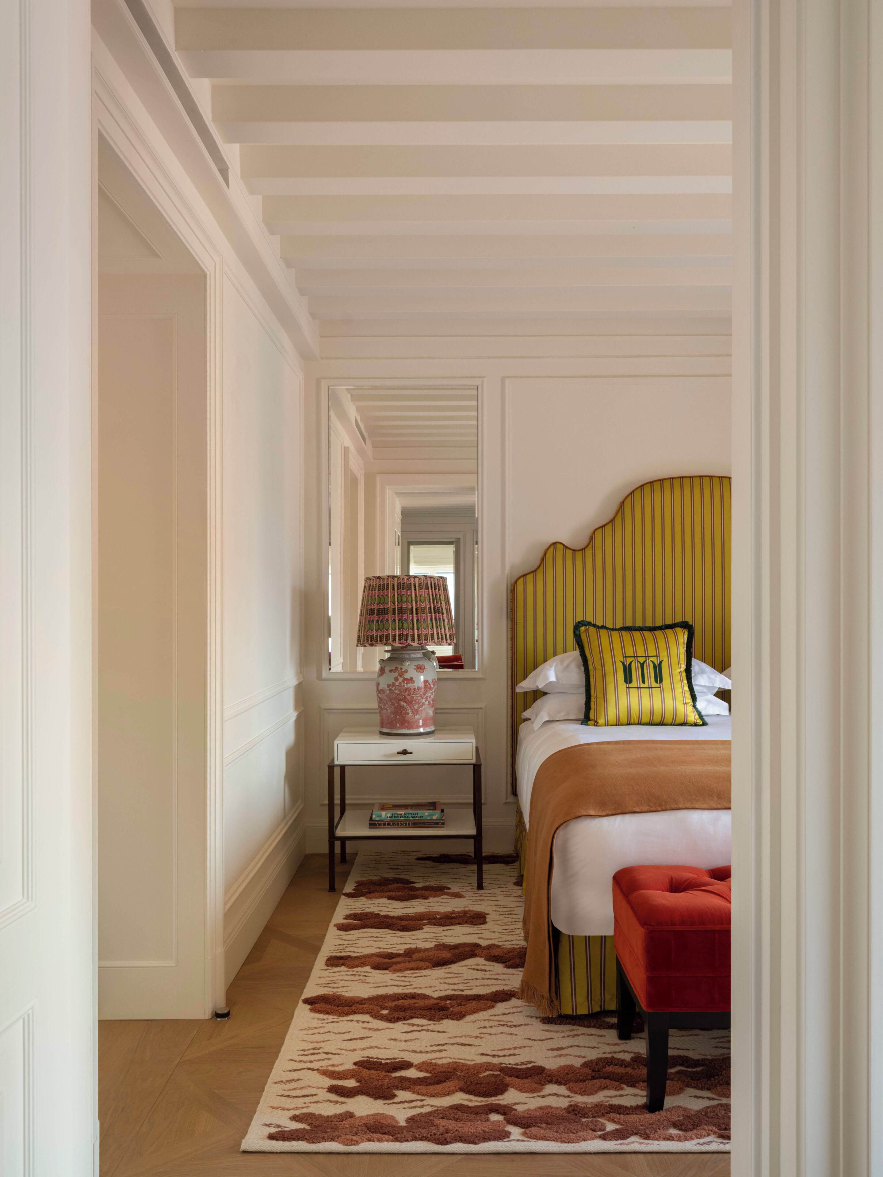 Una habitación de hotel con una cama, una mesita de noche, una lámpara, un banco rojo y una alfombra estampada.