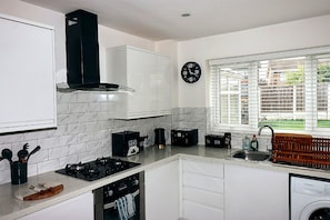 A modern kitchen with white cabinets, a black range hood, a built-in oven, a coffee machine, and a window with blinds. - Cozy West Derby Retreat – Near Alder Hey & Anfield (Liverpool)