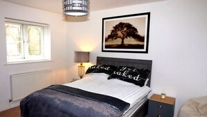 Un dormitorio con cama, lámpara de noche, cuadro enmarco, mesita de noche y ventana con persianas.