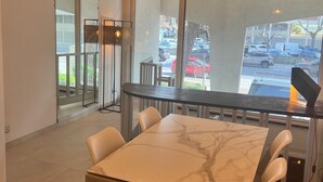 A modern dining area with a marble table and chairs, a hanging light fixture, and a view of the street through large windows. - Les Appartements De Lina - L'Architek (La Grande-Motte)