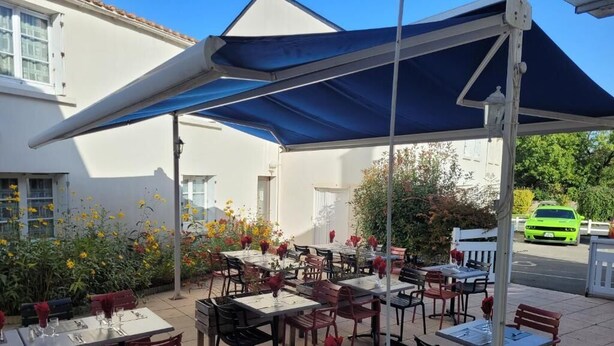 An outdoor dining area with tables and chairs, covered by a blue canopy, and surrounded by flowers and plants. - Le Saint Benoist (Aizenay)