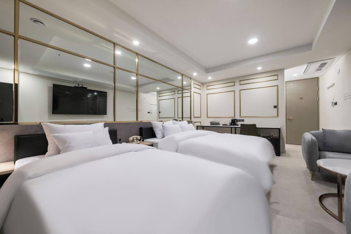 A modern hotel room with two large beds, a desk, a chair, a TV, and a large mirror.