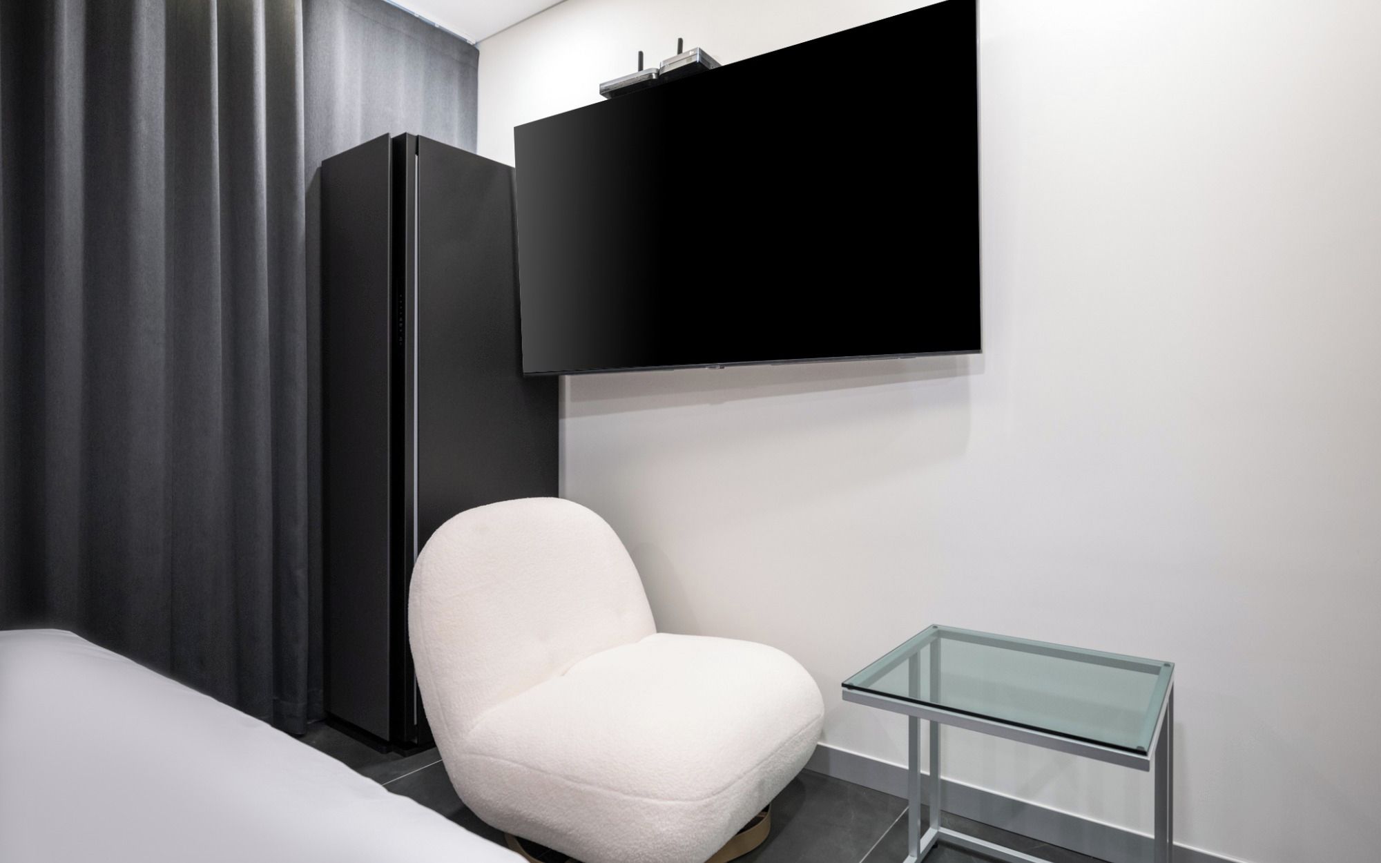 Une chambre d’hôtel moderne avec un lit, un fauteuil blanc, une petite table en verre, un téléviseur noir fixé au mur et une armoire de couleur sombre.
