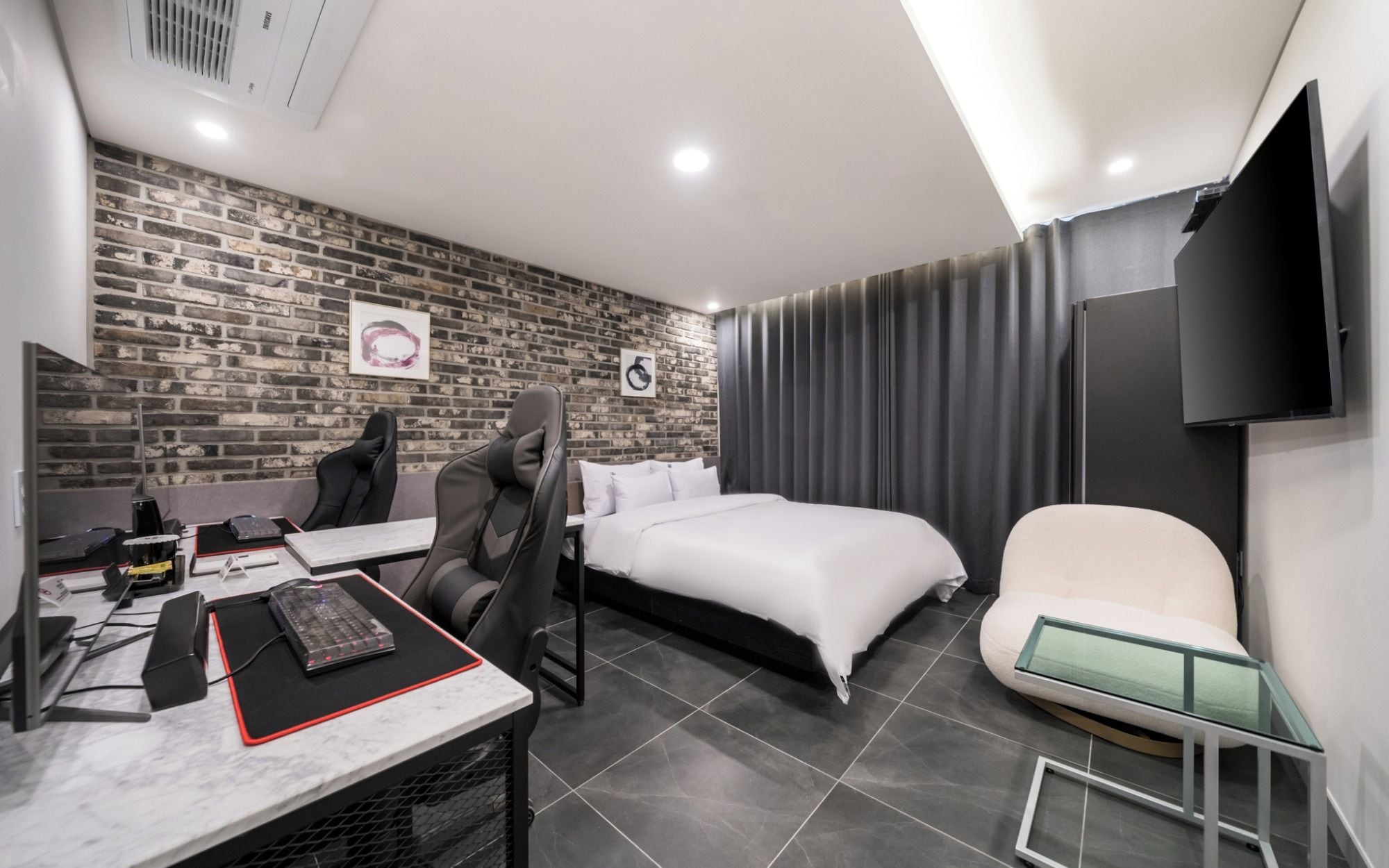A modern hotel room with a brick wall, a bed, a desk with a computer, a chair, and a TV.