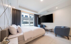 A modern hotel room with a large bed, a TV, a seating area, and a balcony with a view.