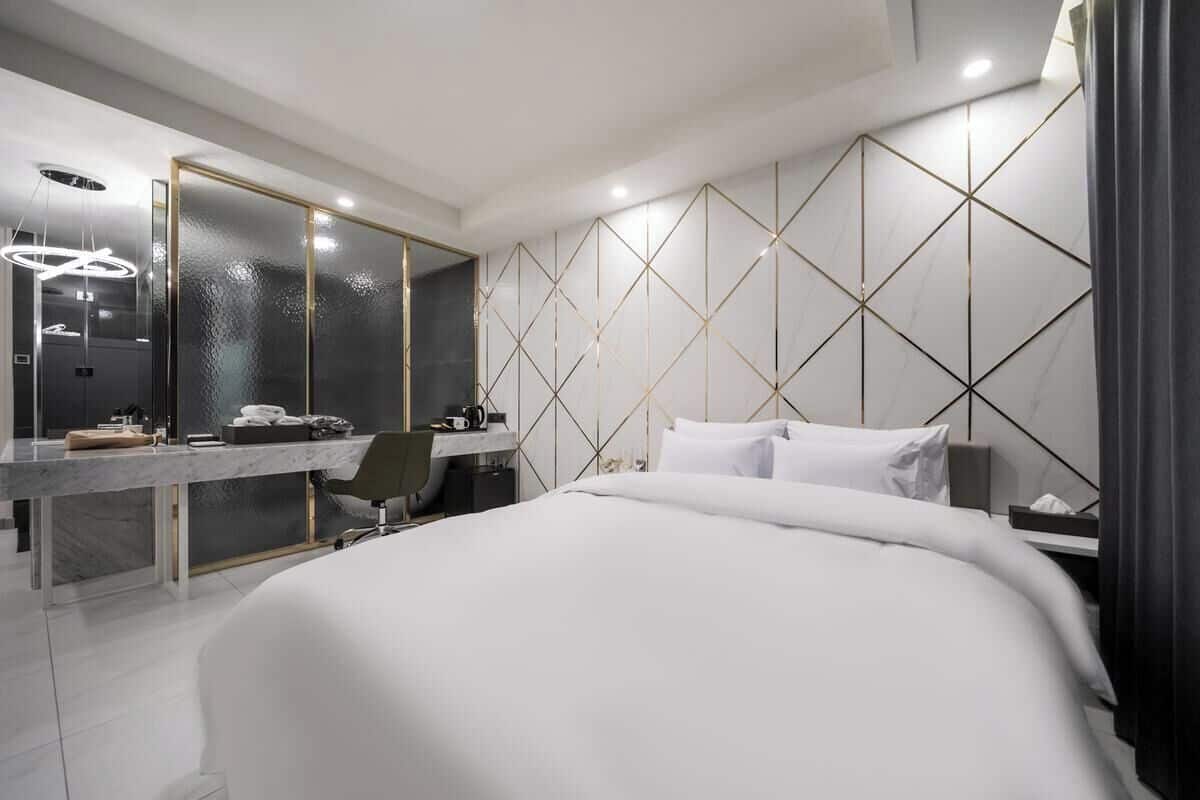 A modern hotel room with a large bed, a desk with a chair, a mirror, and a geometric wall design.