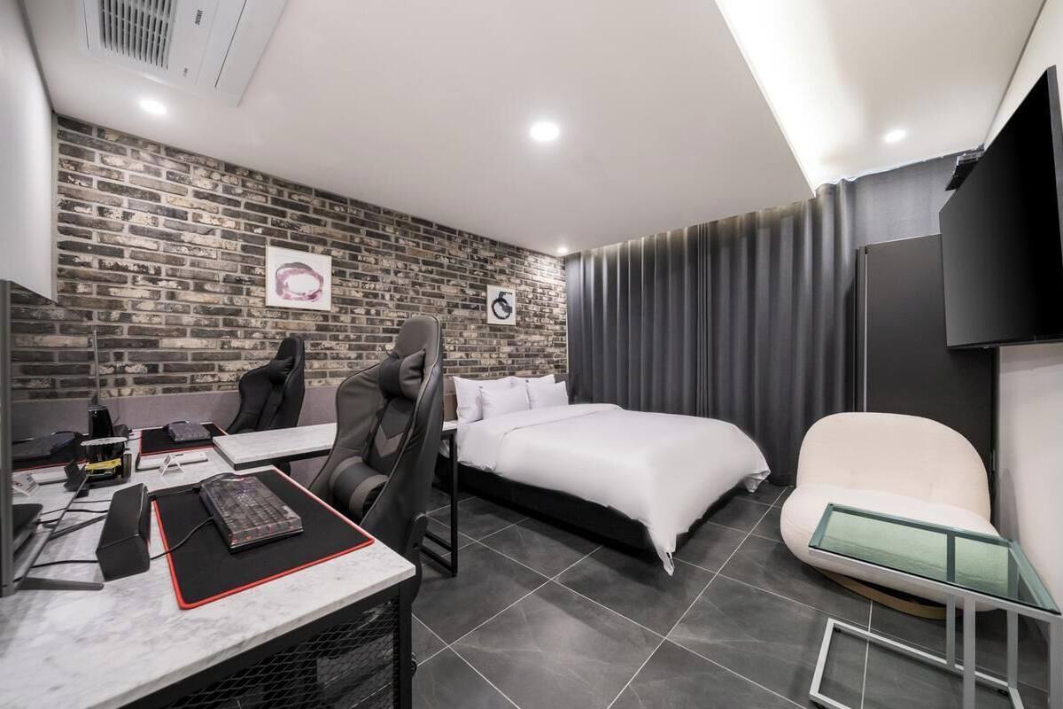 A modern hotel room with a brick-patterned wall, a bed, a desk with a computer, a chair, and a TV mounted on the wall.
