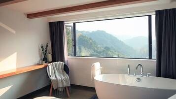 A modern bathroom with a large bathtub, a window with a mountain view, a wooden bench, and a towel rack.