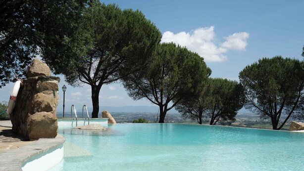 A clear swimming pool with a stone edge, surrounded by trees and a view of distant mountains. - Villa Tiberini Wine Relais (Torrita di Siena)