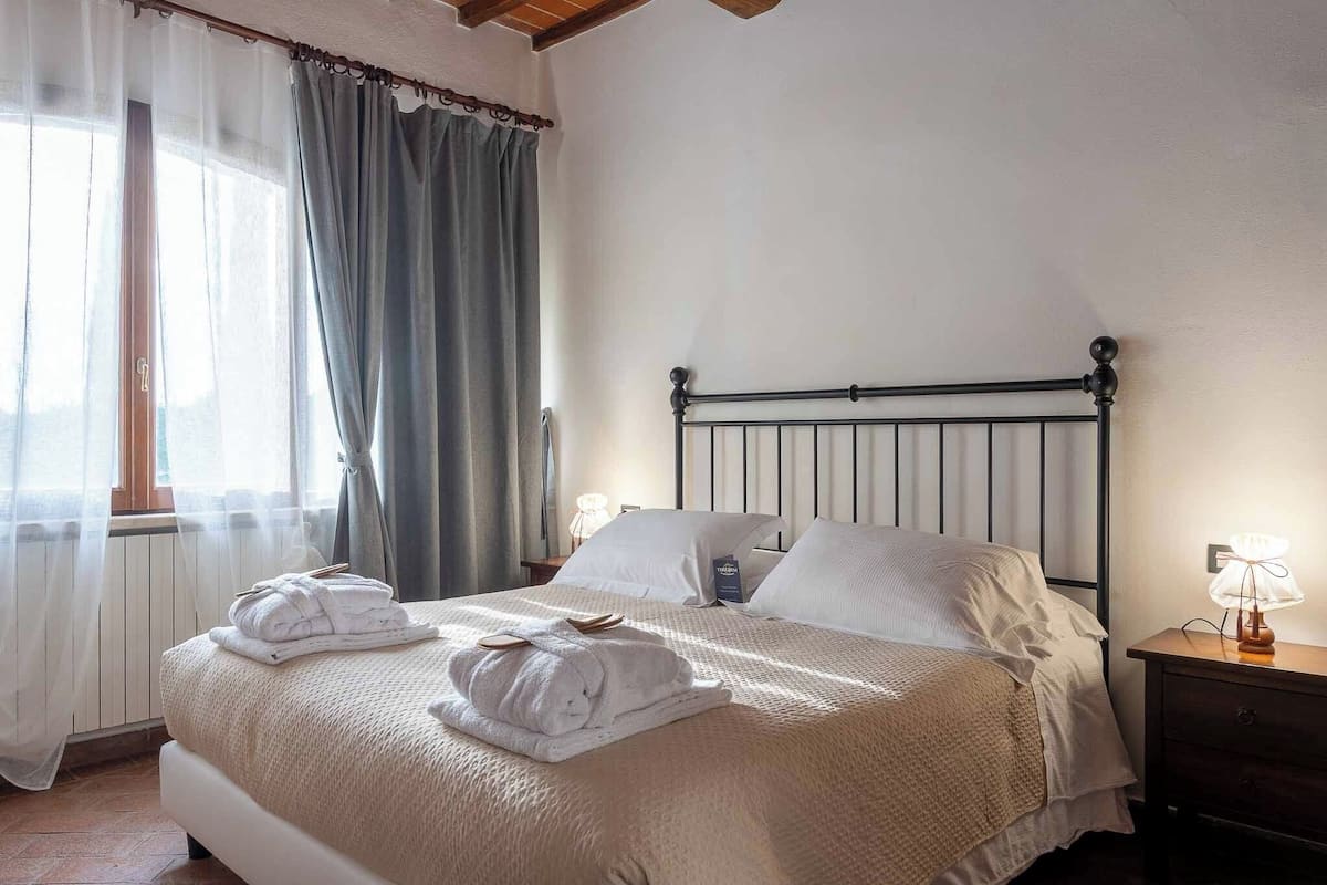 Een net opgemaakt bed met witte lakens en handdoeken, een houten nachtkastje met een lamp, en een raam met dunne gordijnen.
