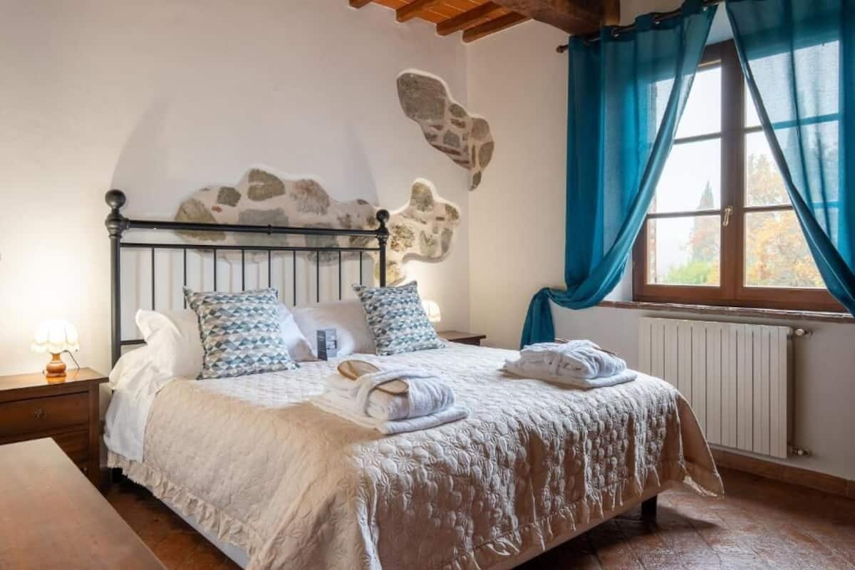 Een net opgemaakt bed met een bekledingspaneel met patroon, een nachtkastje met een lamp en een raam met blauwe gordijnen.