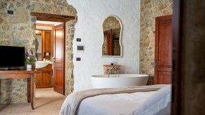 A bedroom with a large bed, a wooden desk, a television, a stone wall, a white bathtub, and a wooden door. - Villa Tiberini Wine Relais (Torrita di Siena)