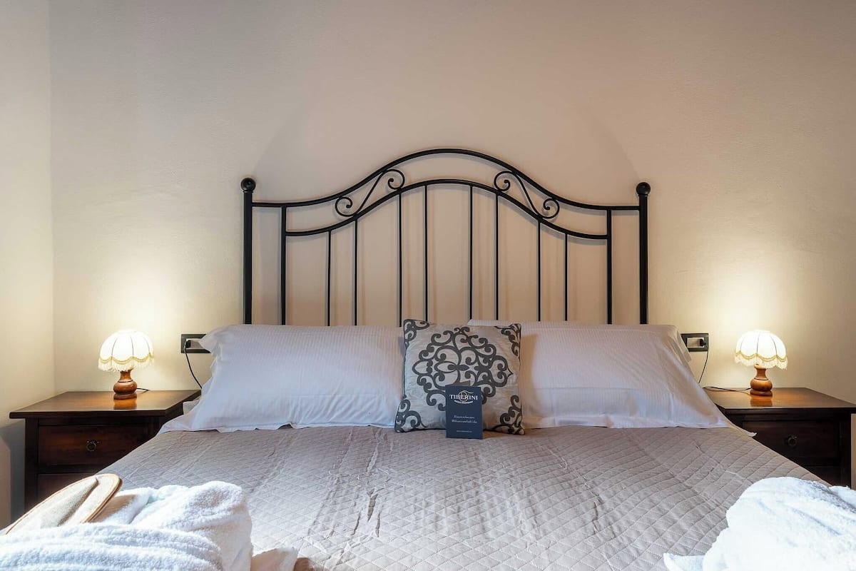 Een net opgemaakt bed met een gewatteerde spreidek, twee bedlampen, een decoratief kussen en een klein bordje op het bed.