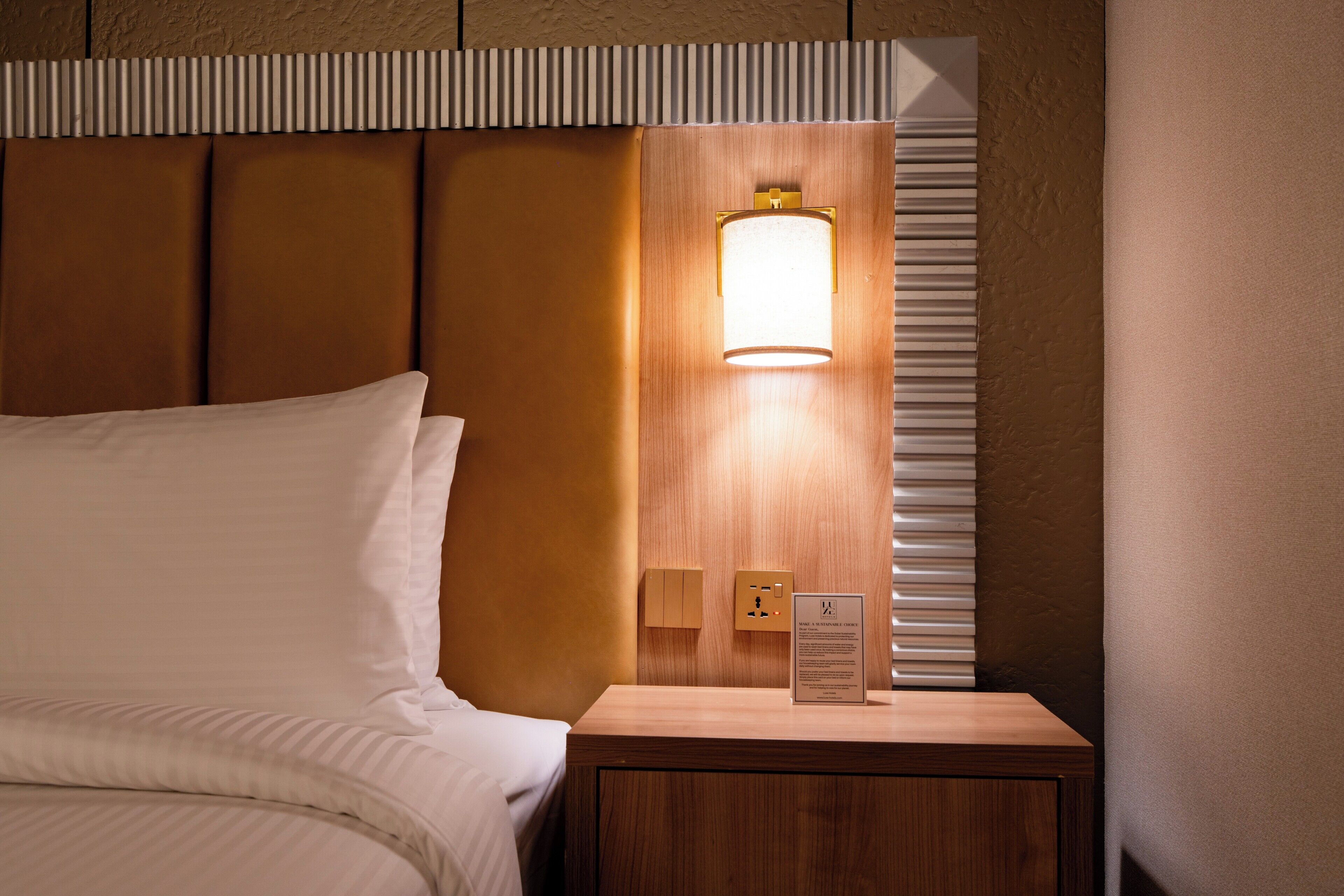 Un letto rifatto con un poggiatesta, un comodino con una lampada e un interruttore a parete.